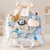 A baby gift basket wrapped in clear cellophane with a large cream satin bow and a small hanging tag. Inside is a coordinated baby gift set including a plush blue and pink animal toy, blue pacifiers, a blue rattle, folded baby clothes, a small plush lion teether, a wooden brush, and a white storage basket with blue stars.